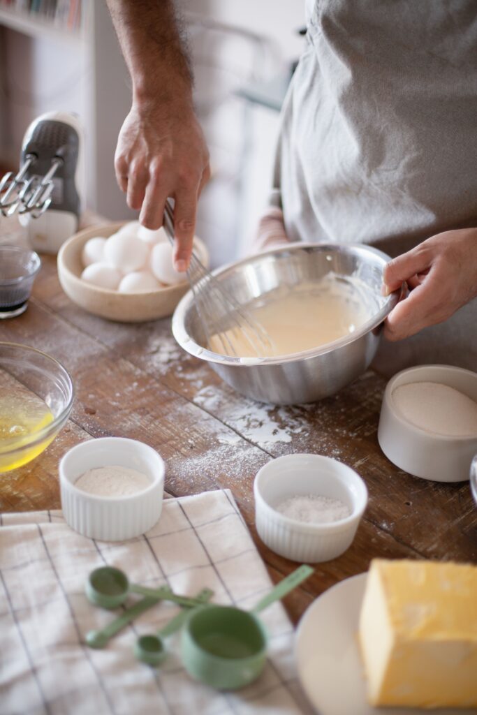Hands whisking soufflé batter in a metal bowl surrounded by eggs, flour, and white ramekins, illustrating the hand-made craft behind a French soufflé