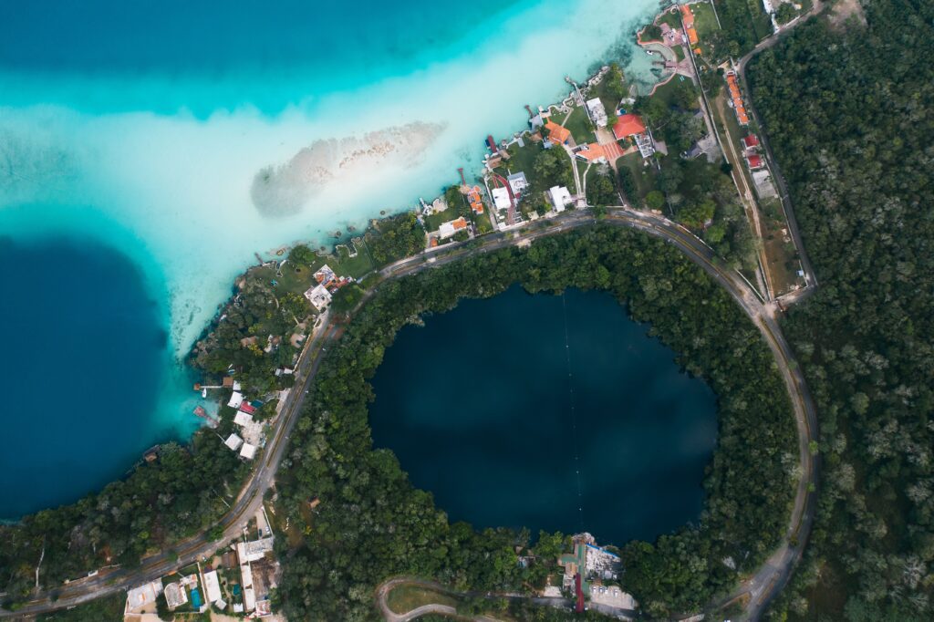 Stunning aerial drone view of Laguna Bacalar Lake of Seven Colors showing turquoise and navy blue water gradients beside a circular cenote surrounded by jungle in Quintana Roo, Mexico