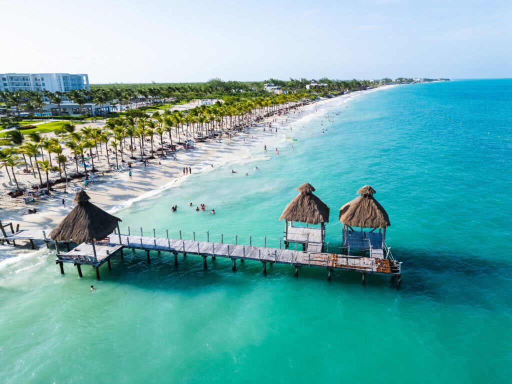 Aerial drone view of a stunning Mexico beach resort with turquoise Caribbean water, wooden palapa pier, white sand, and rows of palm tree
