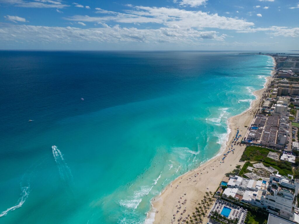 Aerial drone view of Cancún Hotel Zone showing the stunning color gradient from deep navy blue to turquoise Caribbean water along the white sand beach lined with resort hotels in Quintana Roo, Mexico