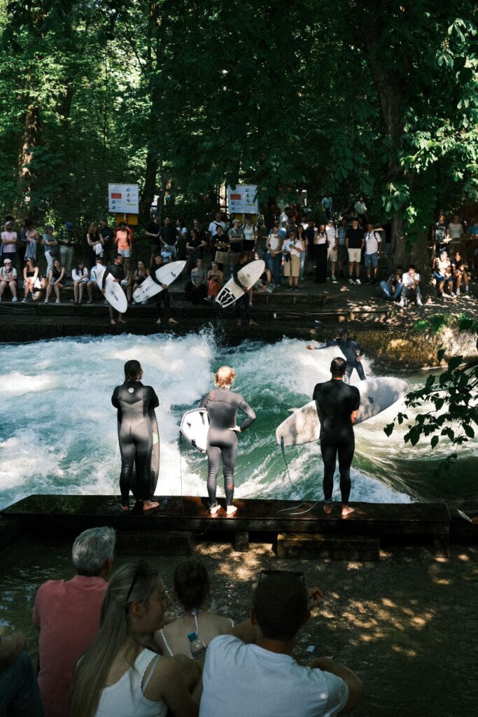 Three surfers in wetsuits holding surfboards standing at the edge of the Eisbach standing wave in the English Garden Munich Germany while a large crowd of spectators watches from the banks