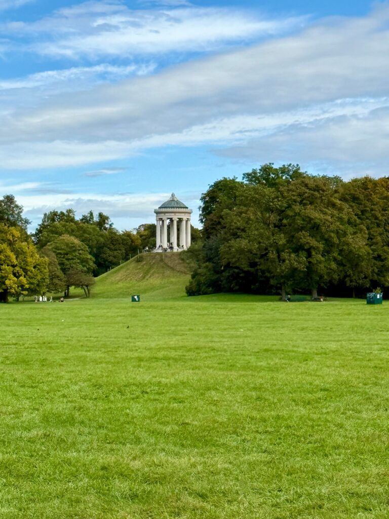The Monopteros Greek temple rotunda on a grassy hill in the English Garden Munich Germany with lush green lawns and autumn trees under a blue sky