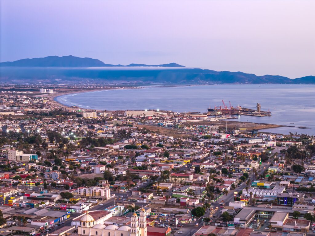 Aerial twilight view of Ensenada, Baja California, Mexico showing the coastal city, commercial port, curved bay and mountain backdrop at dusk