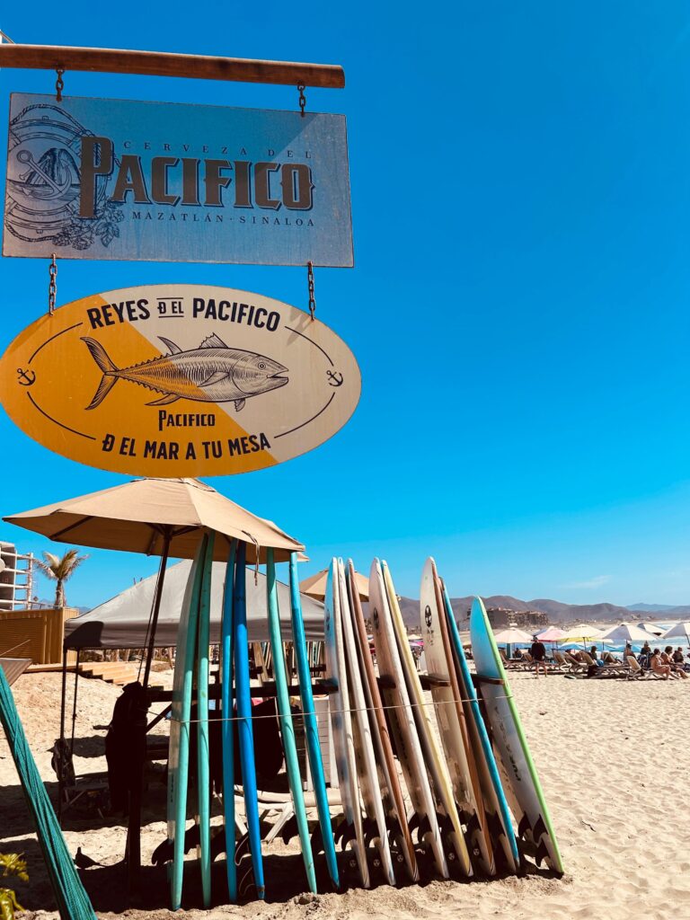 Colorful surfboards lined up for rent on Mazatlán beach beside a Cerveza Pacifico sign with sunbathers and Pacific Ocean mountains visible in the background, Sinaloa, Mexico