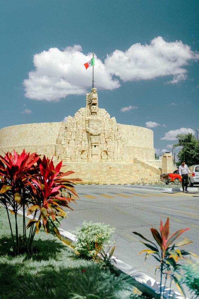 Monumento a la Patria in Mérida, Mexico, with Maya relief carvings and Mexican flag under blue sky