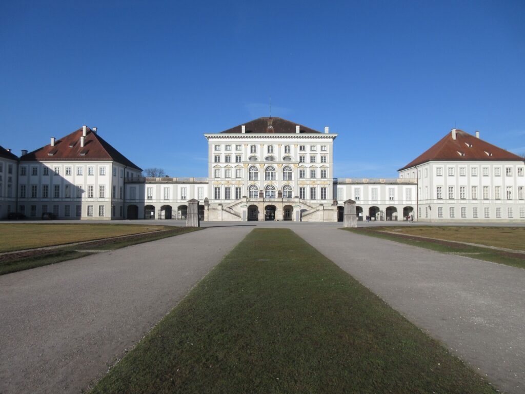 Symmetrical front facade of Nymphenburg Palace Munich Germany showing the grand white Baroque central building flanked by two wing pavilions with a formal grass approach path under a clear blue winter sky
