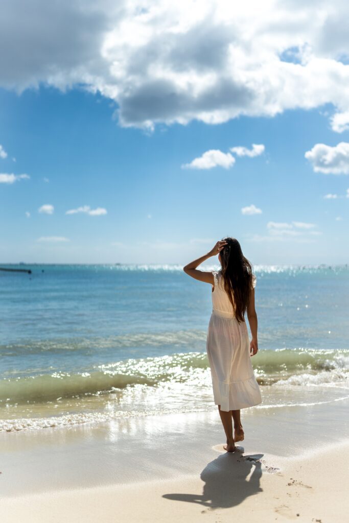 Woman in white dress walking barefoot along the white sand Caribbean beach at Playa del Carmen with calm turquoise water in Quintana Roo, Mexico