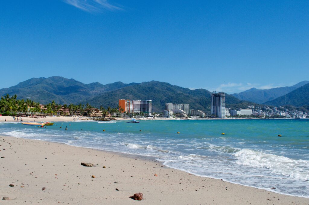 Wide view of Puerto Vallarta beach along Banderas Bay with turquoise Pacific water, colorful beach boats, hotel zone buildings and Sierra Madre mountains in the background, Jalisco, Mexico