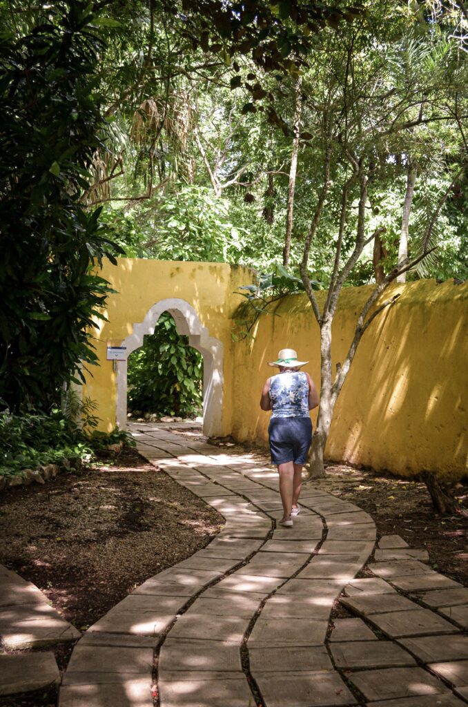 Tourist walking along a brick pathway through a tropical garden with yellow colonial arch wall near Mérida, Yucatán, Mexico