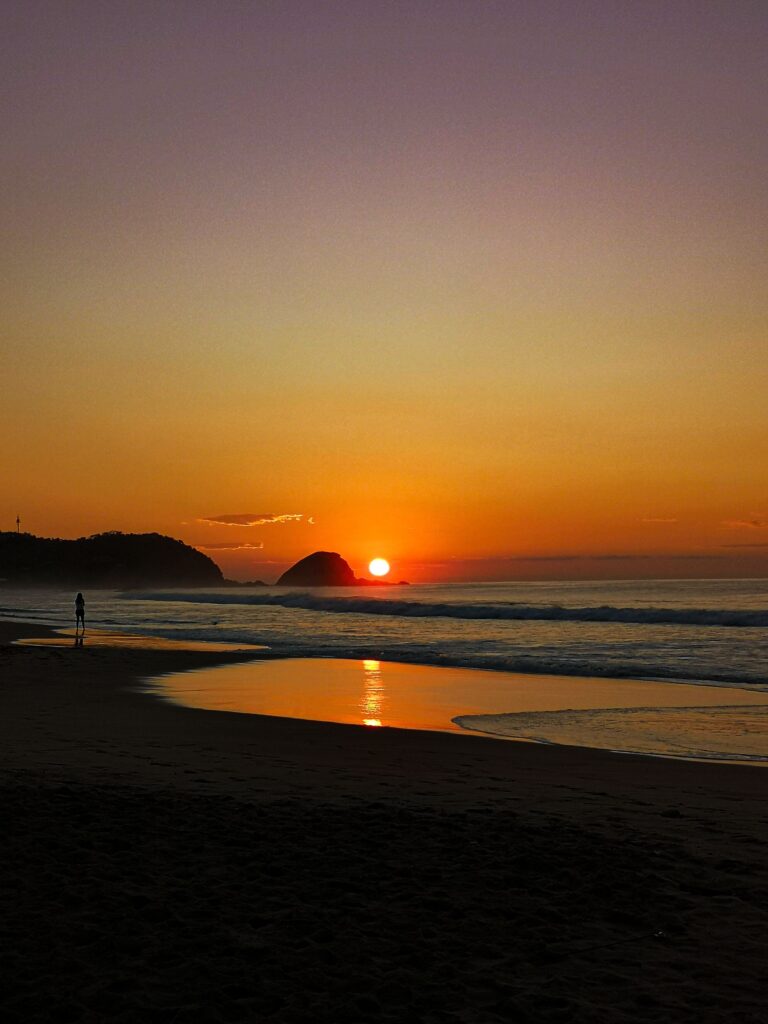 Silhouette of a person standing alone on Zipolite beach at sunset with the sun setting behind a rock formation over the Pacific Ocean in Oaxaca, Mexico